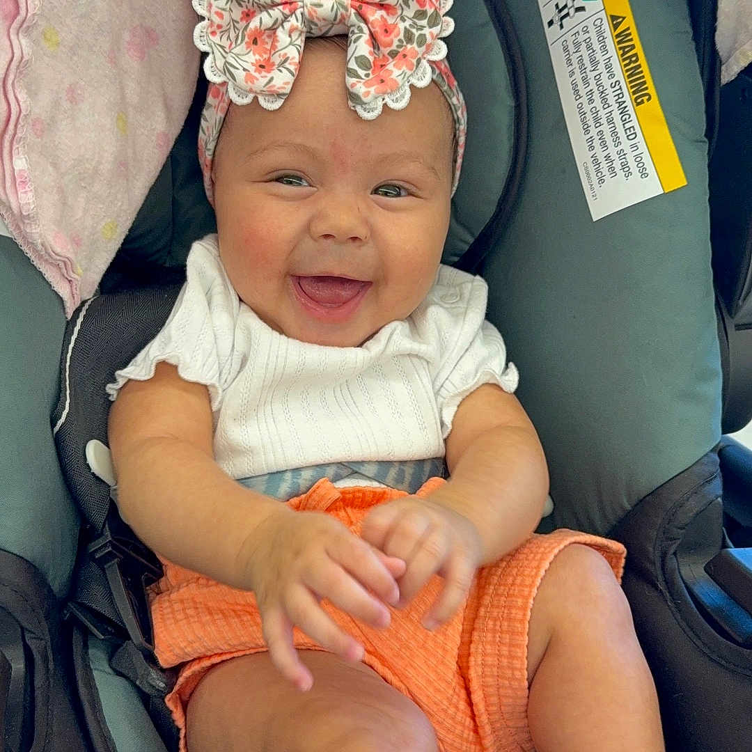 Jaslin joined the competition — help win amazing prizes! baby, blanket, bow, car, car_seat, child, cute, face, hands, happy, headband, indoors, infant, orange_shorts, person, portrait, seatbelt, smiling, socks, white_shirt