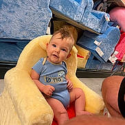 Weston is registered to the contest to win money with this photo: baby, child, plush_chair, blue_chair, store_shelves, floor, person, infant, clothing, short_sleeves, curly_hair, smiling, indoor, retail, soft_texture, seated, looking_at_camera, cute, yellow_chair, playful