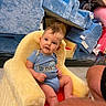 baby, child, plush_chair, blue_chair, store_shelves, floor, person, infant, clothing, short_sleeves, curly_hair, smiling, indoor, retail, soft_texture, seated, looking_at_camera, cute, yellow_chair, playful
