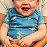 baby, smiling, child, blue_onesie, floral_pattern, hands, adult_hands, holding, happy, portrait, cute, infant, casual_clothing, denim_shorts, beige_shirt, indoor, closeup, person, skin, face