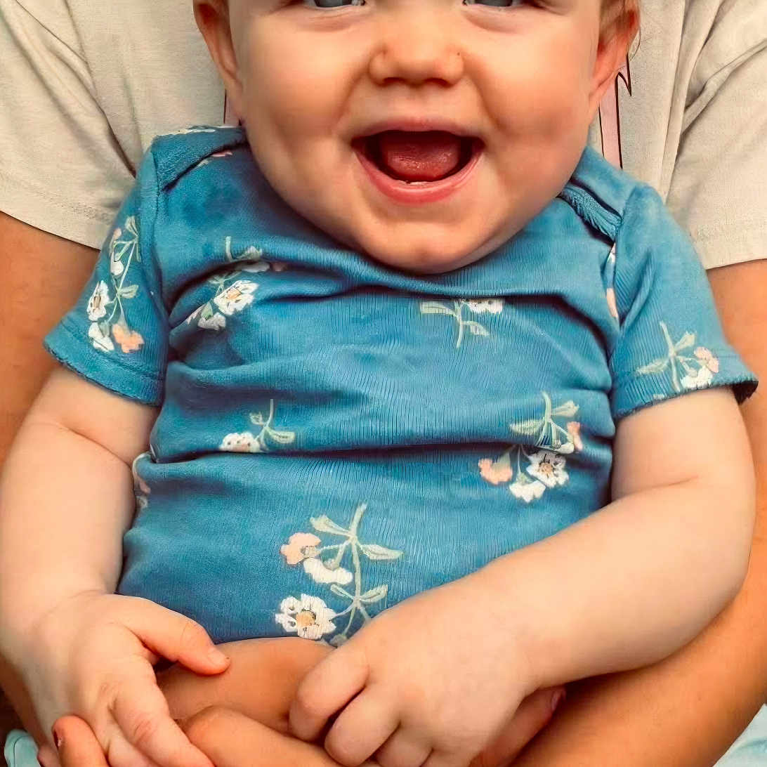 Kleo joined the competition — help win amazing prizes! baby, smiling, child, blue_onesie, floral_pattern, hands, adult_hands, holding, happy, portrait, cute, infant, casual_clothing, denim_shorts, beige_shirt, indoor, closeup, person, skin, face