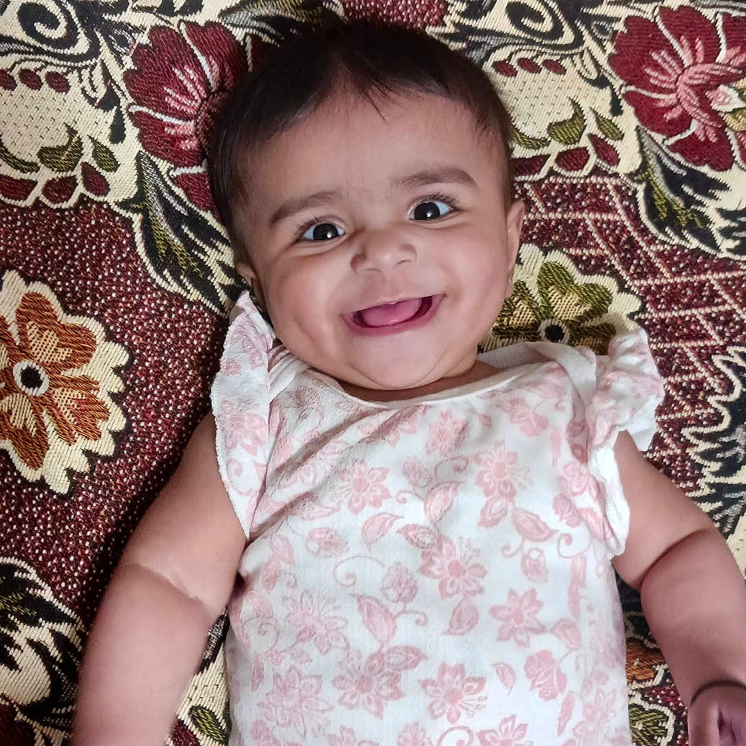 Yashfa is registered to the contest to win money with this photo: baby, smiling, infant, lying_down, onesie, floral_pattern, happy, cute, child, portrait, face, skin, hand, arm, head, fabric, pattern, colorful, background, indoors