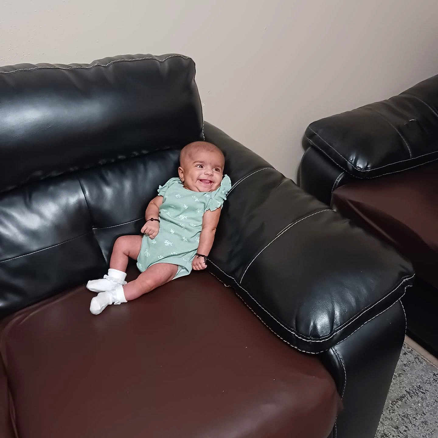 Yashfa is registered to the contest to win money with this photo: baby, infant, smiling, couch, leather, furniture, indoors, socks, onesie, child, happy, person, small, relaxing, living_room, seated, cute, comfortable, home, portrait
