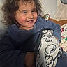 child, smiling, curly_hair, blanket, bed, pillow, indoors, cozy, happy, person, portrait, casual_clothing, blue_sweater, young, cute, resting, comfort, home, relaxed, face