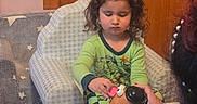 Sebastian is registered to the contest to win money with this photo: child, toddler, toy, mr_potato_head, armchair, pajamas, curly_hair, wooden_wall, table, adult_hand, nail_polish, pearl_hair_accessory, floor, indoor, playing, furniture, person, blue_checkered, green_clothing, star_pattern