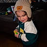 toddler, child, knit_hat, bear_patch, toy, sweater, indoor, living_room, curtain, plush_toys, wooden_floor, sneakers, playful, cute, person, portrait, smiling, young_child, casual_clothing, home