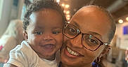 Zephaniah is registered to the contest to win money with this photo: baby, infant, adult, woman, selfie, portrait, smiling, happy, family, 親子, 抱っこ, indoor, warm_lighting, glasses, curly_hair, white_shirt, purple_clothing, football_graphic, restaurant, closeup