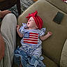 baby, sleeping, couch, red_headband, american_flag, blue_and_white_stripes, infant_clothing, smartphone, adult_leg, gray_sweatpants, wooden_floor, rug, table, hand, furniture, indoor, resting, cute, peaceful, portrait