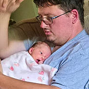 Naomi is registered to the contest to win money with this photo: baby, man, glasses, couch, blanket, indoor, person, holding, newborn, floral_pattern, tattoo, casual_clothing, home, family, affection, portrait, seated, closeup, love, comfort