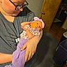 baby, carpet, casual_clothing, eyewear, floral_pattern, glasses, headband, holding, home, human_face, indoor, infant, newborn, parent, person, portrait, purple_clothing, soft_lighting, toddler, wooden_door