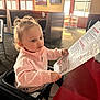 Hadley is registered to the contest to win money with this photo: child, toddler, reading, menu, restaurant, high_chair, pink_jacket, curious, table, menu_paper, portrait, indoor, booth, window, sunlight, hair_tie, hands, seat, face, casual_clothing