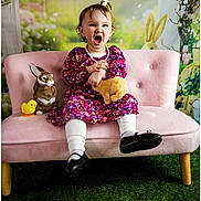 Hadley joined the competition — help win amazing prizes! background, black_shoes, child, cute, decorative_flowers, floral_dress, grass_carpet, happy_expression, indoor, pink_couch, playful, plush_toy, portrait, sitting, smiling, spring_theme, stuffed_bunny, toddler, white_tights, yellow_chick