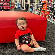 Andres is registered to the contest to win money with this photo: toddler, child, baby, sitting, red_ottoman, carpet, shoe_store, socks, shirt, curly_hair, indoor, footwear, boxes, shelves, retail, store_interior, person, floor, clothing, portrait
