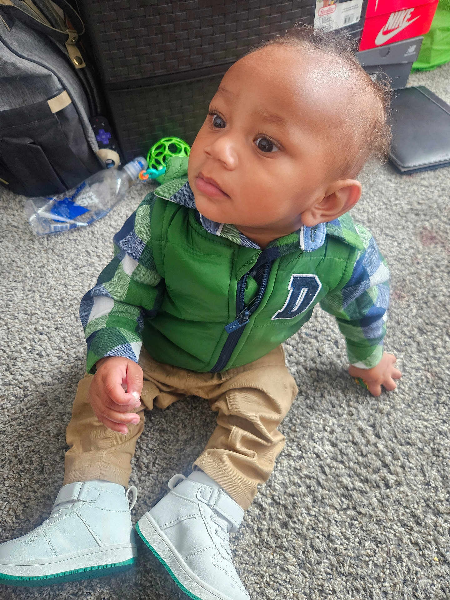 Siyawne joined the competition — help win amazing prizes! backpack, carpet, child, clothing, curious, expression, face, floor, green_vest, hand, home, indoor, person, plaid_shirt, shoe_box, shoes, sitting, toddler, toy, water_bottle