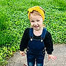 child, toddler, smiling, outdoor, grass, sidewalk, denim_overalls, yellow_headband, happy, person, cute, playful, fashion, skirt, nature, greenery, young, portrait, standing, casual