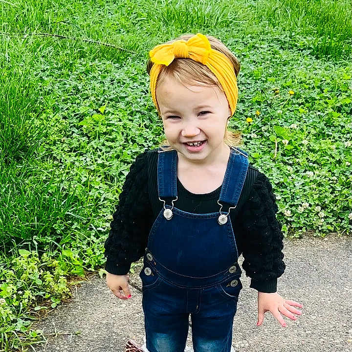 Brooklyn is registered to the contest to win money with this photo: child, toddler, smiling, outdoor, grass, sidewalk, denim_overalls, yellow_headband, happy, person, cute, playful, fashion, skirt, nature, greenery, young, portrait, standing, casual