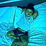 barefoot, bed, blue_light, calm, casual_clothing, child, comfort, furniture, home, indoor, person, pillow, portrait, relaxing, resting, sheet, shorts, sleepwear, tshirt, young_boy