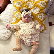 Eliana joined the competition — help win amazing prizes! baby, infant, couch, pillow, headband, socks, dress, patterned_pillow, beige_couch, child, cute, indoors, furniture, textile, fabric, portrait, happy, small_child, sitting, cozy