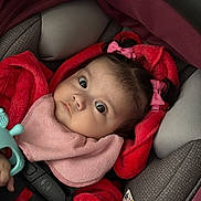 Eliana is registered to the contest to win money with this photo: baby, child, car_seat, red_clothing, pink_bow, toy, infant, wide_eyes, cute, hand, blanket, seatbelt, indoor, portrait, closeup, face, curious, soft_toys, warm_clothing, young_child