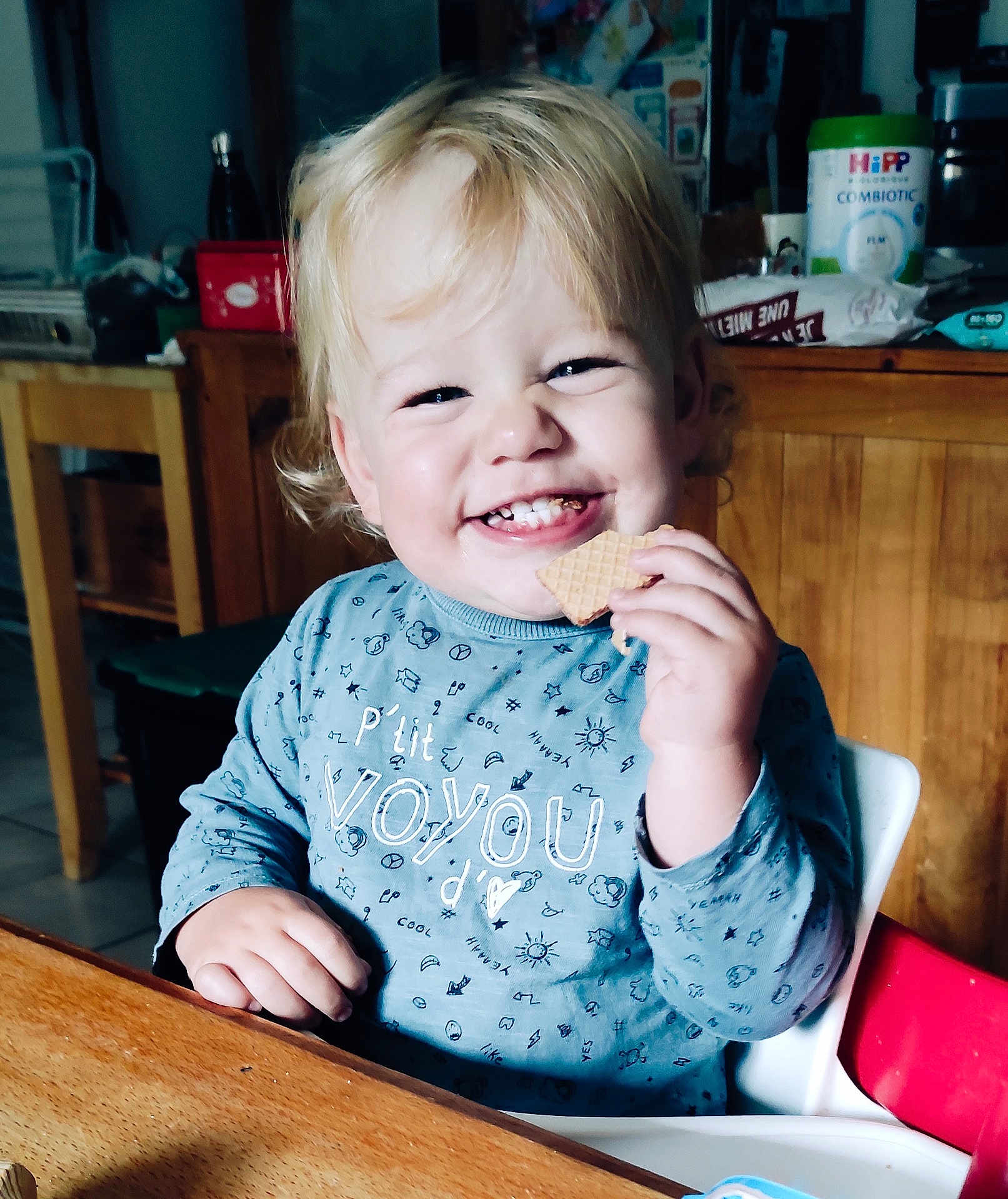 Samuel a rejoint le concours — aidez-le/la à gagner de superbes lots ! toddler, child, smiling, cookie, high_chair, blonde_hair, blue_shirt, kitchen, wooden_table, happy, indoor, person, food, snack, cute, sitting, young_child, face, portrait, meal