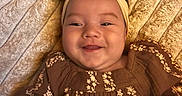 Leilani is registered to the contest to win money with this photo: baby, child, infant, smiling, portrait, lying_down, headband, bow, floral_dress, brown_clothing, blanket, bedding, close_up, face, eyes, happy, indoors, soft_texture, warm_lighting, photography