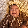 baby, child, infant, smiling, portrait, lying_down, headband, bow, floral_dress, brown_clothing, blanket, bedding, close_up, face, eyes, happy, indoors, soft_texture, warm_lighting, photography