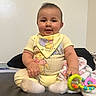baby, child, infant, sitting, portrait, smiling, face, yellow_outfit, bib, toys, rattle, bed, blanket, pillow, indoors, bedroom, home, cute, looking_at_camera, baby_toy