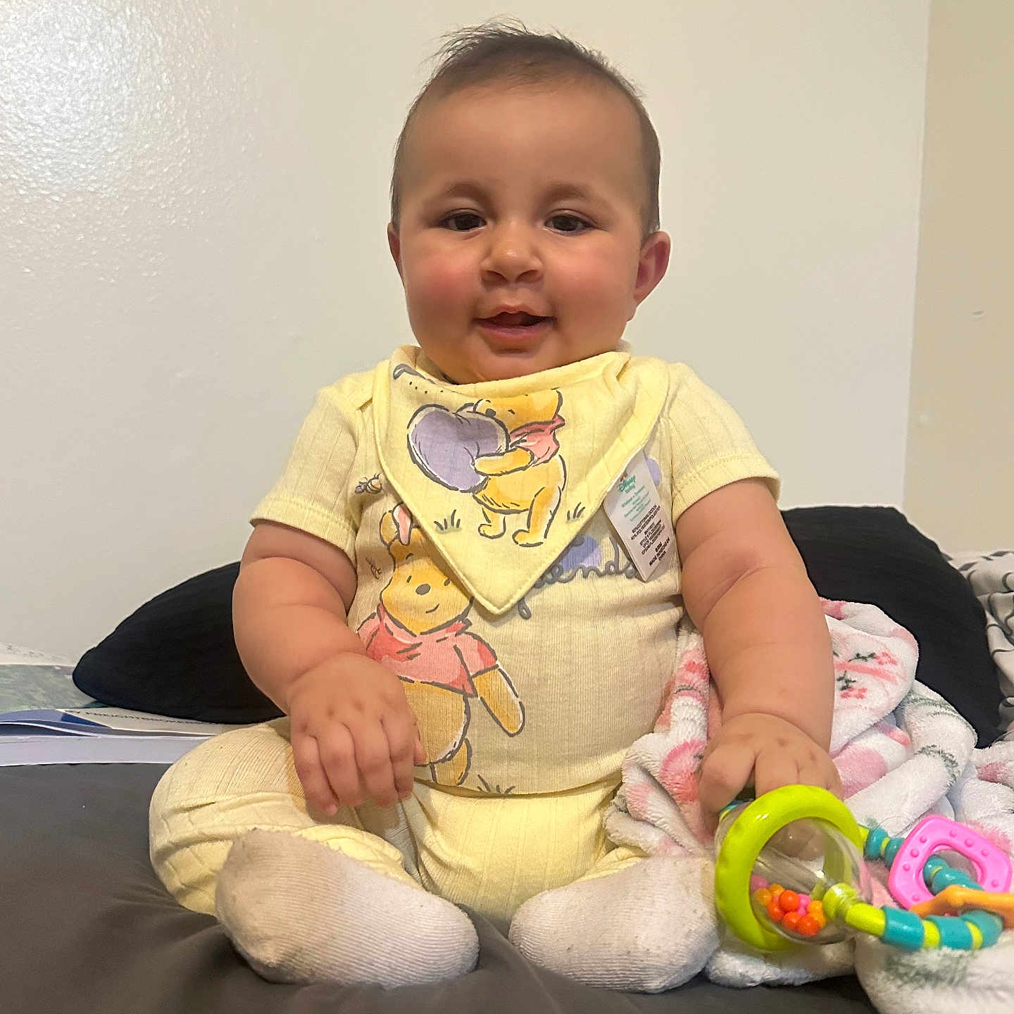 Zenlayha is registered to the contest to win money with this photo: baby, child, infant, sitting, portrait, smiling, face, yellow_outfit, bib, toys, rattle, bed, blanket, pillow, indoors, bedroom, home, cute, looking_at_camera, baby_toy