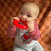 Elijah is registered to the contest to win money with this photo: baby, child, toy, red, striped_clothing, gray_pants, chewing, sitting, indoor, cute, smiling, face, hands, playful, background, fabric, infant, happy, closeup, portrait