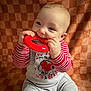 baby, child, toy, red, striped_clothing, gray_pants, chewing, sitting, indoor, cute, smiling, face, hands, playful, background, fabric, infant, happy, closeup, portrait