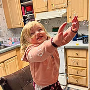 Bella is registered to the contest to win money with this photo: child, toddler, bubble, kitchen, hoodie, smiling, blonde_hair, indoor, cabinet, dishwasher, countertop, chair, playful, happy, reaching, floral_pants, home, cute, person, fun