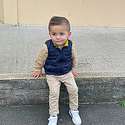 Timéo participe au concours pour gagner de l'argent avec cette photo : child, boy, sitting, concrete, sidewalk, wall, casual_clothing, white_shoes, vest, curly_hair, outdoor, young, portrait, fashion, street, cute, casual, curious, daylight, urban