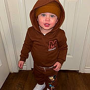 Myles is registered to the contest to win money with this photo: toddler, child, brown_hoodie, beanie, mickey_mouse, white_shoes, indoor, wooden_floor, smiling, standing, blue_eyes, face, clothing, hood_up, pants, casual_wear, person, door, floor, cute