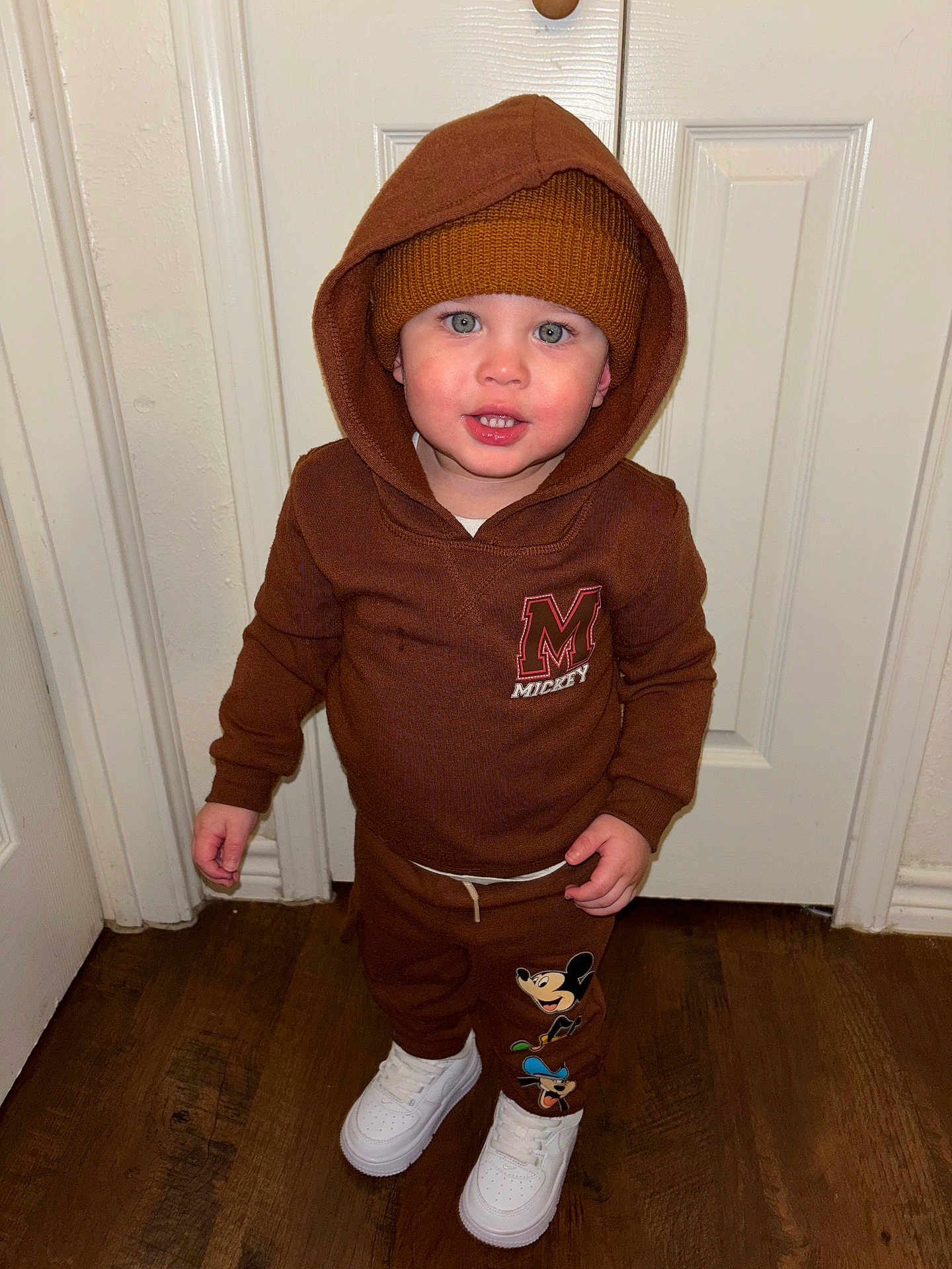 Myles is registered to the contest to win money with this photo: toddler, child, brown_hoodie, beanie, mickey_mouse, white_shoes, indoor, wooden_floor, smiling, standing, blue_eyes, face, clothing, hood_up, pants, casual_wear, person, door, floor, cute