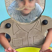 Tiago participe au concours pour gagner de l'argent avec cette photo : child, kid, face, playground, play_equipment, scratched_plastic, mustache_sticker, sticker, hands, portrait, curious, patterned_shirt, denim_shorts, sand, outdoor, blue_panel, yellow_surface, window_cutout, black_knob, closeup