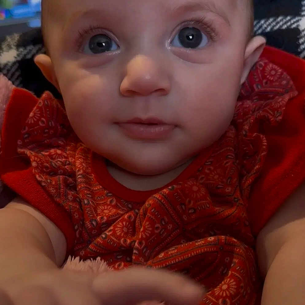 Kyrsten is registered to the contest to win money with this photo: baby, child, closeup, curious, cute, expression, eyes, face, furniture, hand, indoor, infant, patterned_outfit, portrait, red_clothing, sitting, skin, soft_blanket, warm_lighting, young_child