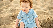 Colton is registered to the contest to win money with this photo: child, toddler, curly_hair, blue_outfit, sand, beach, sitting, smiling, outdoor, sunlight, cute, young_child, portrait, playful, summer, skin, face, hands, legs, nature
