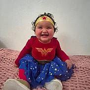 Kylie is registered to the contest to win money with this photo: baby, child, costume, wonder_woman, headband, smiling, sitting, indoor, blanket, knitted, red_clothing, blue_skirt, gold_star, curly_hair, white_shoes, cute, portrait, happy, person, young