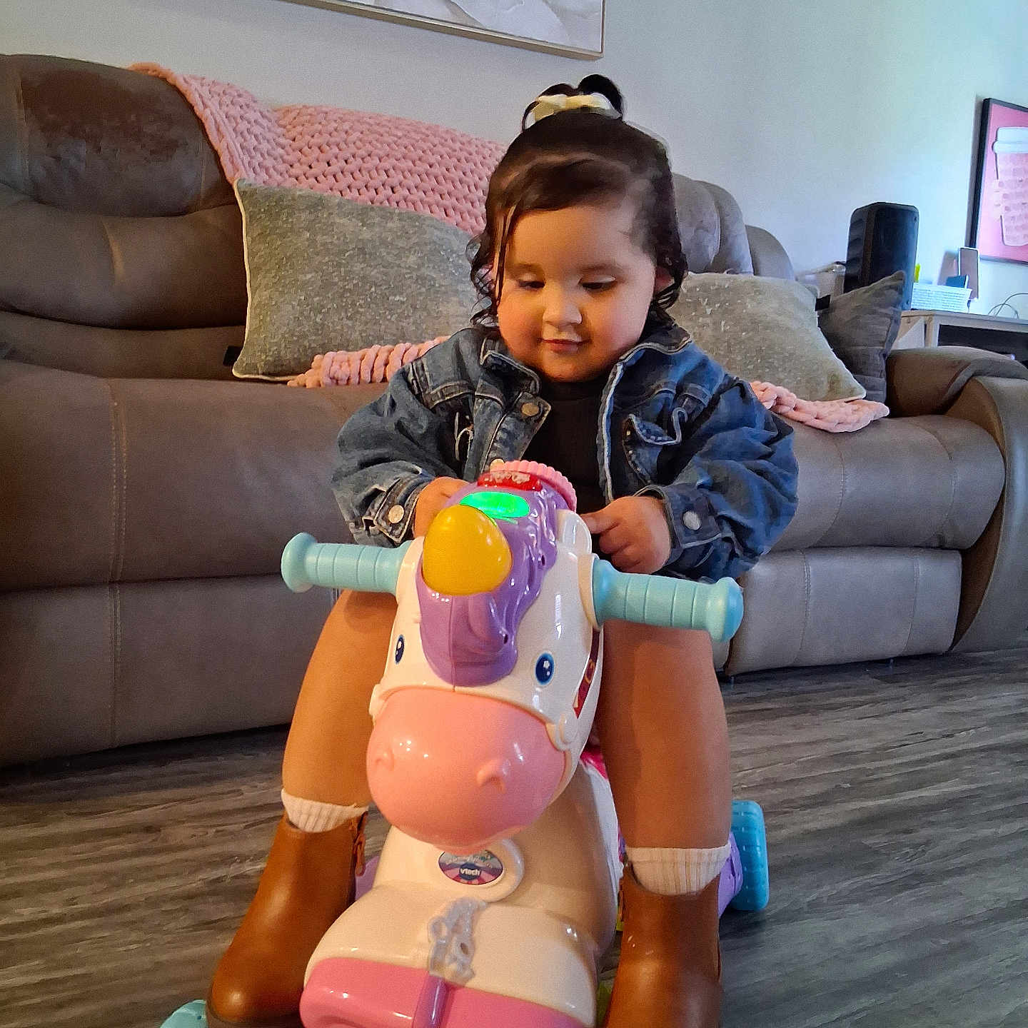 Kylie is registered to the contest to win money with this photo: toddler, child, toy, unicorn, ride_on_toy, denim_jacket, brown_boots, indoor, couch, pillow, blanket, painting, flooring, happy, cute, playful, person, furniture, home, smile