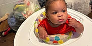 Brylen is registered to the contest to win money with this photo: baby, child, infant, red_shirt, face, hand, floating_ring, indoor, floor, plastic_bag, clothing, person, wooden_floor, curly_hair, expression, seat, toy, household_items, colorful, looking_away