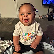 Brylen is registered to the contest to win money with this photo: baby, smiling, happy, child, toddler, indoor, clothing, shirt, blanket, carpet, living_room, person, cute, sitting, expression, furniture, television, shorts, skin, joy