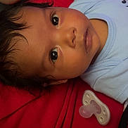 Rumani joined the competition — help win amazing prizes! baby, infant, child, portrait, lying_down, wide_eyes, dark_hair, blue_clothing, red_blanket, pacifier, plush_toy, close_up, indoors, cozy, face, head, eyes, bedding, soft_lighting, photography