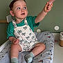 toddler, child, baby, overalls, socks, bottle, changing_pad, indoor, green_wall, polka_dots, curious, sitting, cute, clothing, hand, face, expression, furniture, room, person