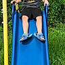 child, boy, slide, playground, outdoor, grass, greenery, batman_tshirt, blue_slide, yellow_pole, smiling, shorts, footwear, shark_slippers, happy, summer, nature, fun, play, leisure