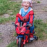 child, boy, smiling, toy_motorcycle, spiderman_costume, bandana, outdoor, grass, gravel_path, happy, playful, costume, footwear, toy, riding, summer, fun, casual, nature, portrait