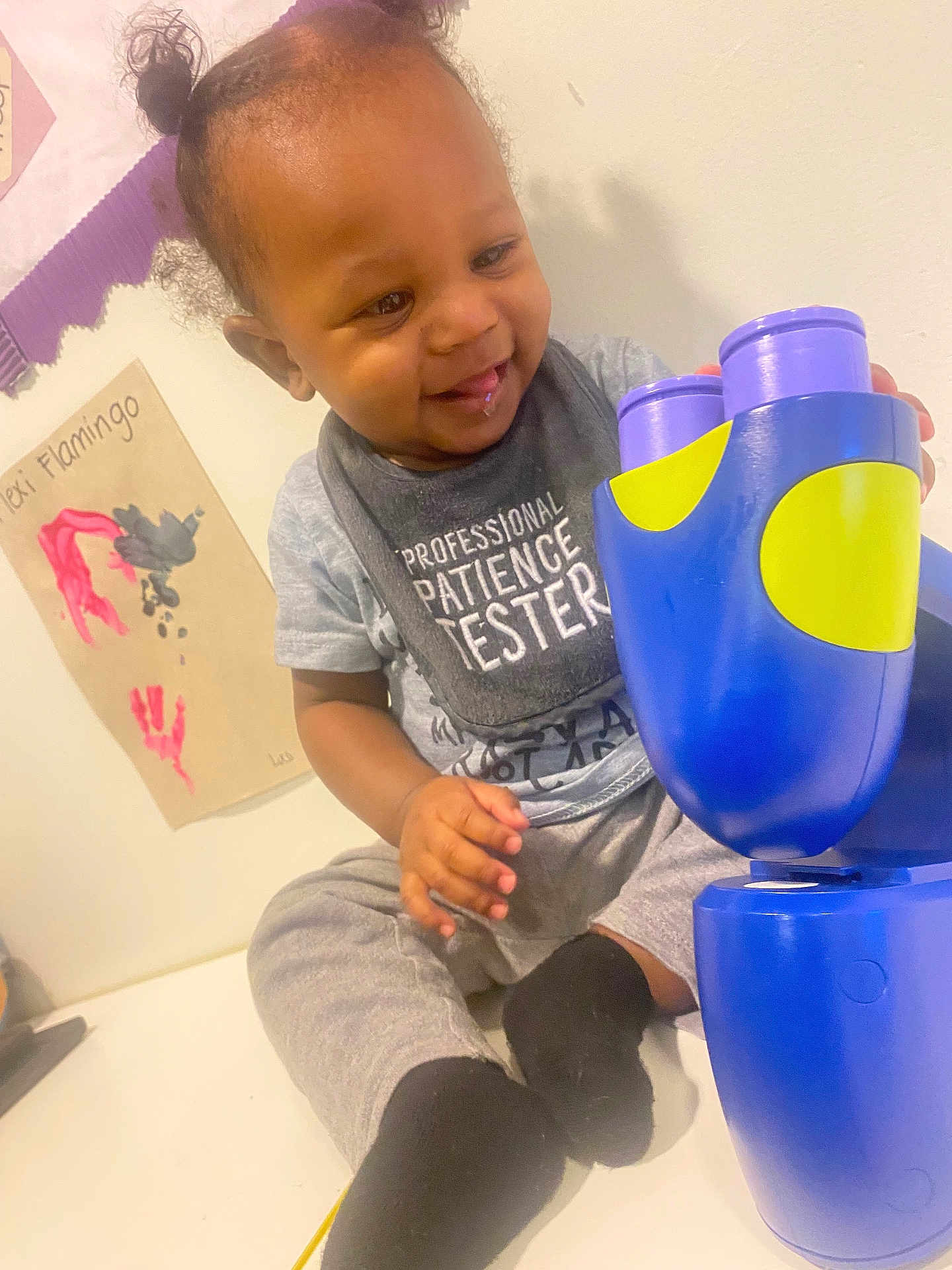 Karsyn is registered to the contest to win money with this photo: toddler, child, smiling, indoor, toy, bib, clothing, black_socks, hair_pigtails, face, hand, sitting, table, wall_art, flamingo, play, happy, casual_wear, person, portrait