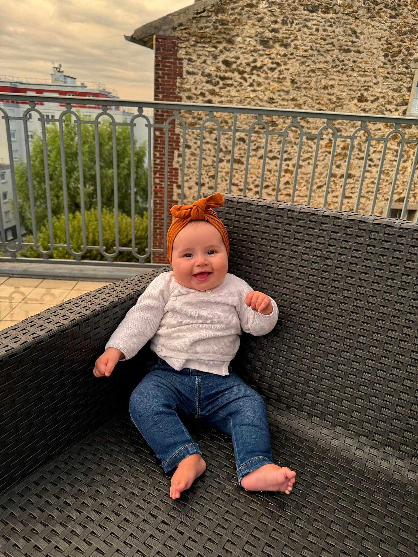 Saja a rejoint le concours — aidez-le/la à gagner de superbes lots ! baby, child, smiling, outdoor, chair, woven_chair, headband, orange_headband, jeans, white_cardigan, barefoot, railing, stone_wall, building, sky, greenery, portrait, happy, cute, sitting