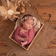 Ella participe au concours pour gagner de l'argent avec cette photo : newborn, baby, sleeping, wooden_box, floral_headband, wrapped, burlap, dried_flowers, flower_arrangement, soft_cloth, peaceful, indoors, portrait, infant, cute, resting, natural_light, decor, wood_floor, cozy