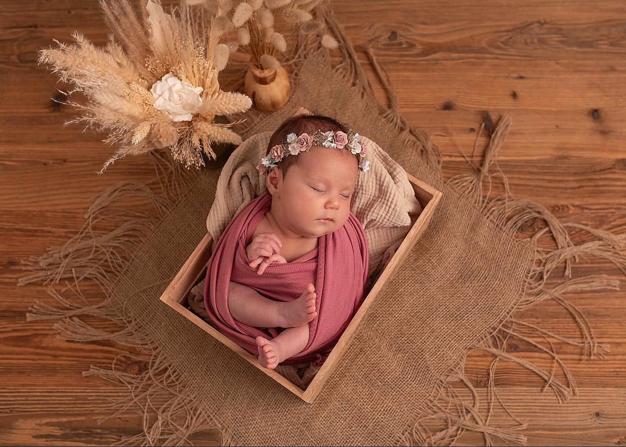 Ella participe au concours pour gagner de l'argent avec cette photo : newborn, baby, sleeping, wooden_box, floral_headband, wrapped, burlap, dried_flowers, flower_arrangement, soft_cloth, peaceful, indoors, portrait, infant, cute, resting, natural_light, decor, wood_floor, cozy