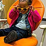 child, young_child, smiling, yellow_sunglasses, braided_hair, pink_jacket, denim_jacket, orange_chair, dental_chair, dentist_office, sitting, playful, happy, black_shoes, indoor, portrait, accessory, hand_up, cute, shoe_sole